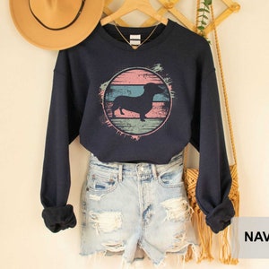 May include: A navy blue cropped sweatshirt with a vintage-style graphic of a dachshund silhouette in a pink, blue, and green circle. The sweatshirt is worn over a pair of light blue denim shorts.