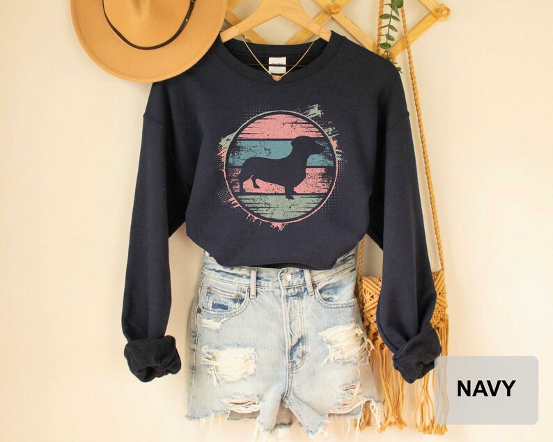 May include: A navy blue cropped sweatshirt with a vintage-style graphic of a dachshund silhouette in a pink, blue, and green circle. The sweatshirt is worn over a pair of light blue denim shorts.