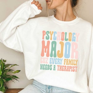 May include: White sweatshirt with the text "Psychology Major Cuz Every Family Needs A Therapist" in rainbow colours.