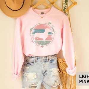 May include: Light pink sweatshirt with a vintage-style graphic of a dachshund in a circle. The graphic has a faded, distressed look with shades of pink, blue, and green.