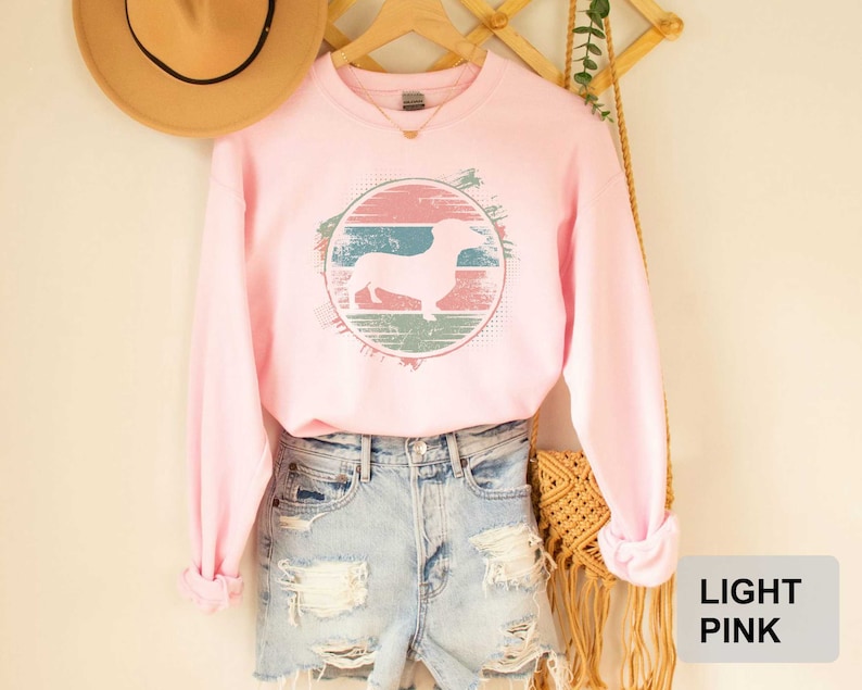 May include: Light pink sweatshirt with a vintage-style graphic of a dachshund in a circle. The graphic has a faded, distressed look with shades of pink, blue, and green.