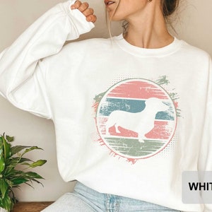 May include: White sweatshirt with a vintage-style graphic of a dachshund in a circle with pink, blue, and green stripes. The text "WHITE" is visible in the bottom right corner of the image.