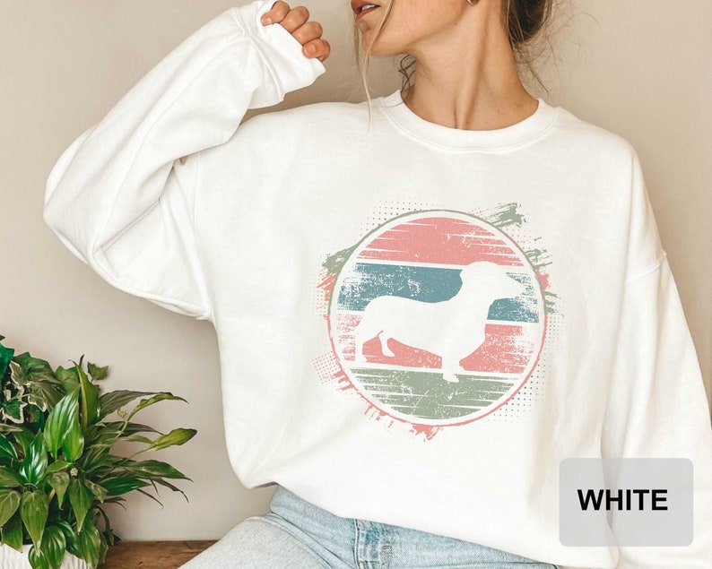 May include: White sweatshirt with a vintage-style graphic of a dachshund in a circle with pink, blue, and green stripes. The text "WHITE" is visible in the bottom right corner of the image.