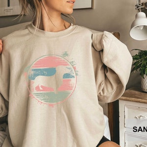 May include: A light beige sweatshirt with a graphic print of a dachshund silhouette in a colorful circle with a retro sunset design. The text "SAND" is visible in the lower right corner of the image.