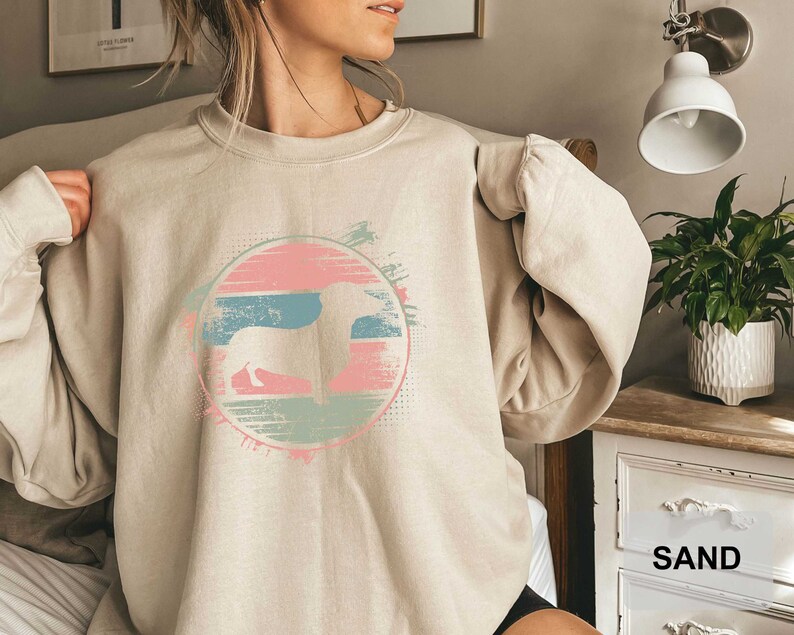 May include: A light beige sweatshirt with a graphic print of a dachshund silhouette in a colorful circle with a retro sunset design. The text "SAND" is visible in the lower right corner of the image.