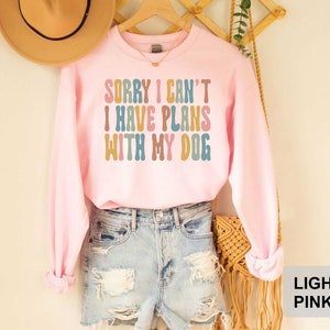 May include: A light pink sweatshirt with the text "Sorry I can't I have plans with my dog" in colorful retro font.