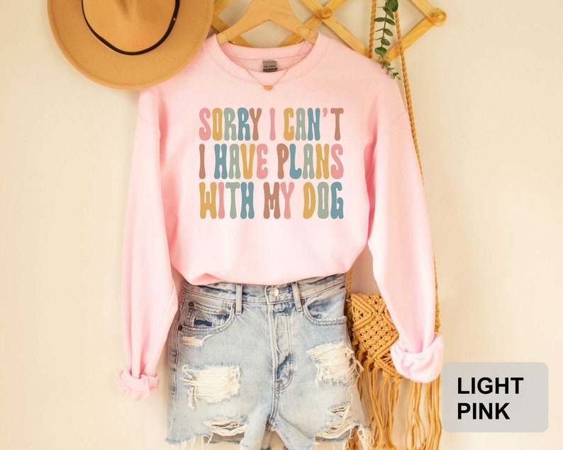 May include: A light pink sweatshirt with the text "Sorry I can't I have plans with my dog" in colorful retro font.