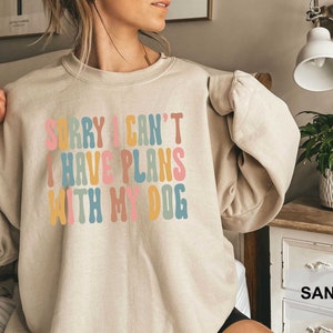 May include: A light beige sweatshirt with the text "SORRY I CAN'T I HAVE PLANS WITH MY DOG" in colorful, rounded letters.