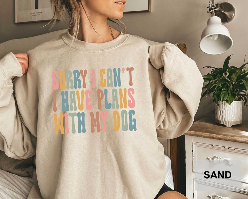 May include: A light beige sweatshirt with the text "SORRY I CAN'T I HAVE PLANS WITH MY DOG" in colorful, rounded letters.