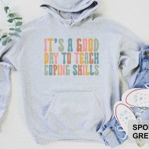 May include: Sport grey hoodie with the text "IT'S A GOOD DAY TO TEACH COPING SKILLS" in retro-style lettering. The hoodie has a front pocket and a drawstring hood. The hoodie is paired with blue jeans and white sneakers.