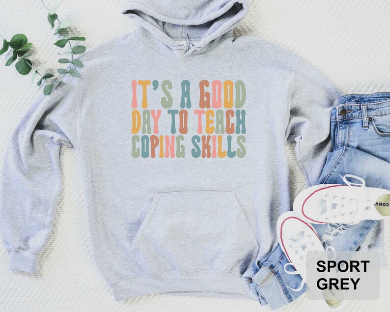 May include: Sport grey hoodie with the text "IT'S A GOOD DAY TO TEACH COPING SKILLS" in retro-style lettering. The hoodie has a front pocket and a drawstring hood. The hoodie is paired with blue jeans and white sneakers.