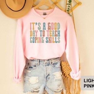 May include: Light pink sweatshirt with the text "IT'S A GOOD DAY TO TEACH COPING SKILLS" in colorful, retro-style lettering. The long-sleeved top is paired with denim shorts and a straw hat.