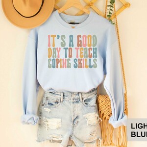May include: Light blue sweatshirt with the text "IT'S A GOOD DAY TO TEACH COPING SKILLS" in colorful retro font. The sweatshirt is displayed on a wooden hanger with a straw hat and denim shorts in the background.
