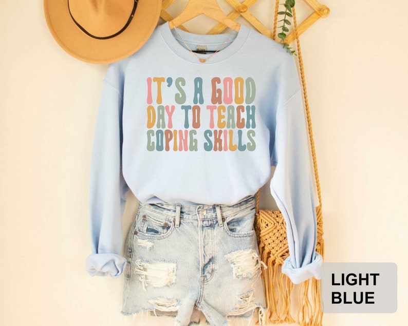 May include: Light blue sweatshirt with the text "IT'S A GOOD DAY TO TEACH COPING SKILLS" in colorful retro font. The sweatshirt is displayed on a wooden hanger with a straw hat and denim shorts in the background.