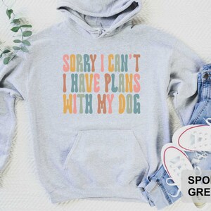 May include: A gray hooded sweatshirt with the text "Sorry I can't I have plans with my dog" in colorful, retro-style lettering. The sweatshirt is shown on a white background with blue jeans and white sneakers.