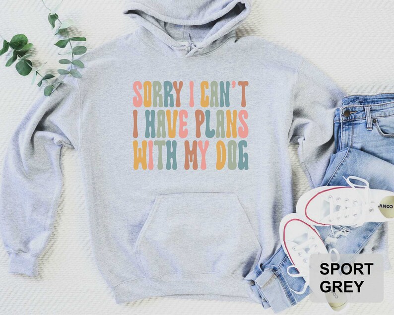 May include: A gray hooded sweatshirt with the text "Sorry I can't I have plans with my dog" in colorful, retro-style lettering. The sweatshirt is shown on a white background with blue jeans and white sneakers.