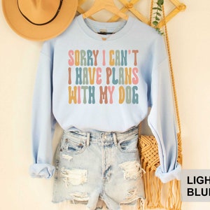 May include: Light blue sweatshirt with the text "Sorry I can't I have plans with my dog" in colorful retro lettering.