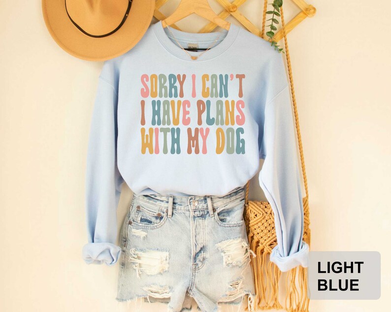 May include: Light blue sweatshirt with the text "Sorry I can't I have plans with my dog" in colorful retro lettering.