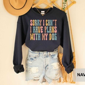 May include: Navy blue cropped sweatshirt with the text "Sorry I can't I have plans with my dog" in rainbow colors. The sweatshirt is worn with denim shorts and a brown fringed bag.