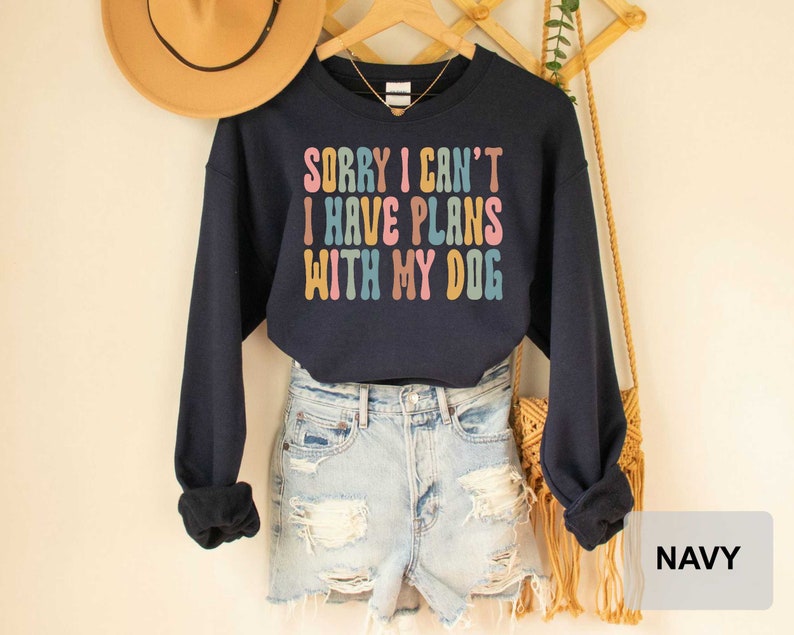 May include: Navy blue cropped sweatshirt with the text "Sorry I can't I have plans with my dog" in rainbow colors. The sweatshirt is worn with denim shorts and a brown fringed bag.