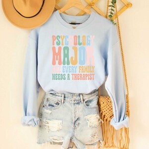 May include: Light blue sweatshirt with the text "Psychology Major cuz every family needs a therapist" in rainbow colours.