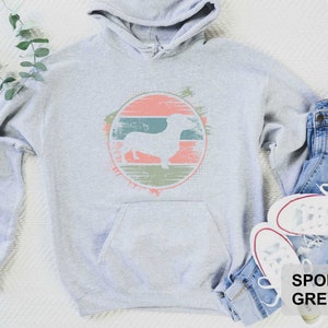 May include: A gray hooded sweatshirt with a vintage-style graphic of a dachshund in a circle. The graphic has a peach, green, and brown color scheme. The text "SPORT GREY" is printed on the bottom right corner of the image.