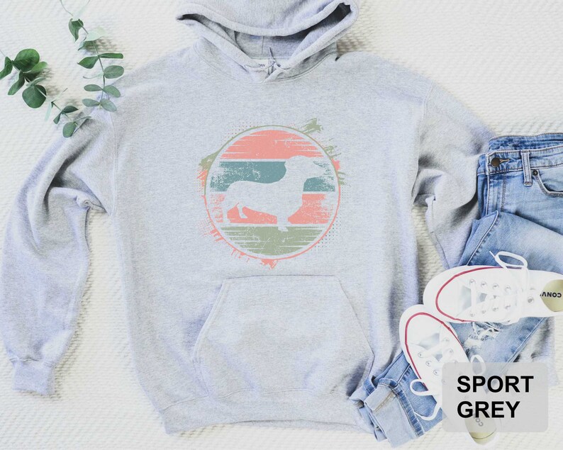 May include: A gray hooded sweatshirt with a vintage-style graphic of a dachshund in a circle. The graphic has a peach, green, and brown color scheme. The text "SPORT GREY" is printed on the bottom right corner of the image.