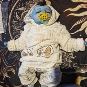May include: A blue-skinned doll with dark hair and closed eyes, wearing a cream-colored quilted hooded jacket with 'Disney THE LION KING' text and a Simba graphic, paired with matching white pants featuring a repeating Simba print and a yellow pacifier.