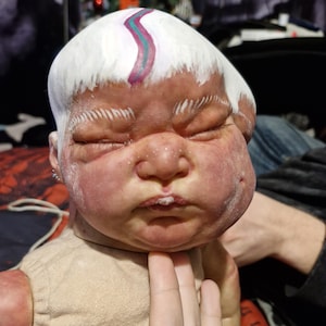 May include: A realistic baby doll with a white painted head and a pink and blue stripe painted on the forehead. The doll is wearing a beige onesie.