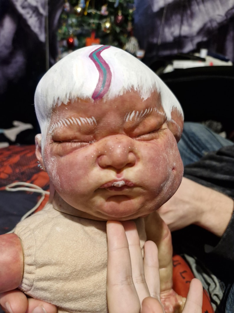 May include: A realistic baby doll with a white painted head and a pink and blue stripe painted on the forehead. The doll is wearing a beige onesie.