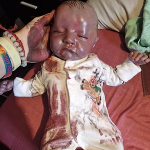 Könnte beinhalten: Eine Reborn-Puppe mit geschlossenen Augen, die einen weißen Strampler mit einem Rentier-Motiv trägt. Der Strampler hat ein rotes und graues Muster. Die Haut der Puppe hat eine dunkle, purpurrote Farbe.