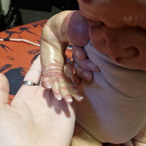 May include: A close-up of a realistic baby doll with a light brown complexion and a white onesie. The doll's arm is outstretched and the hand is open. The doll's face is turned away from the camera.