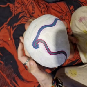 May include: A white, round object with a blue and purple swirl design painted on it. The object is being held by a person's hand.