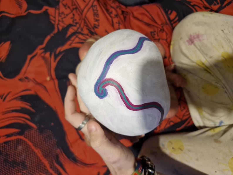 May include: A white, round object with a blue and purple swirl design painted on it. The object is being held by a person's hand.