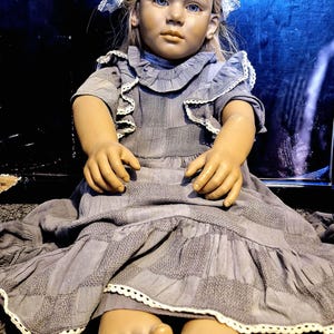 May include: A porcelain doll with blonde hair wearing a gray dress with white lace trim. The doll has blue eyes and is sitting on a brown carpet.