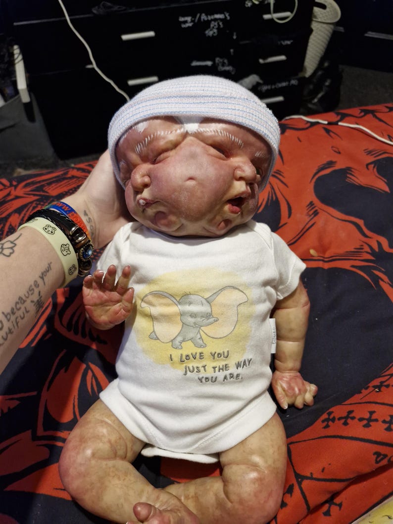 May include: A realistic baby doll with two faces, wearing a white onesie with an elephant graphic and the text "I love you just the way you are."