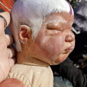 May include: A close-up of a doll's head with white paint and a purple stripe. The doll has a realistic face with closed eyes and a slightly open mouth. The doll's hair is styled in a short, straight cut.