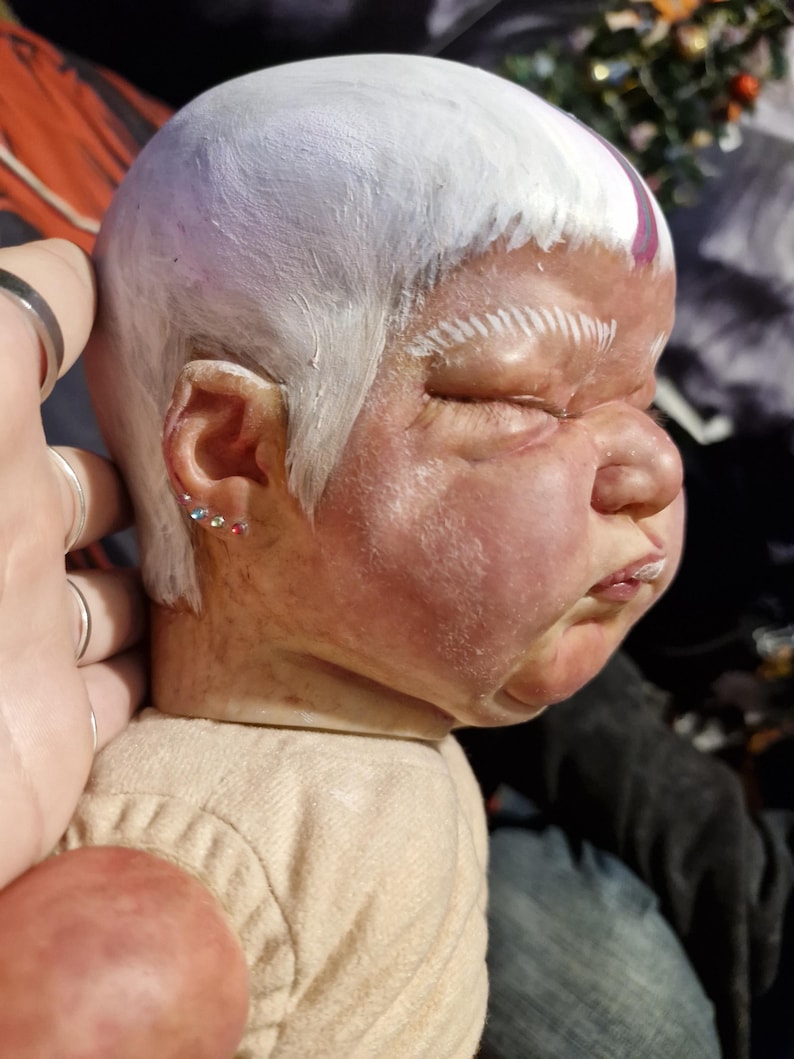 May include: A close-up of a doll's head with white paint and a purple stripe. The doll has a realistic face with closed eyes and a slightly open mouth. The doll's hair is styled in a short, straight cut.