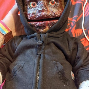 May include: A creepy doll wearing a black hoodie with a zipper. The doll has a realistic face with red eyes and a bloody appearance.
