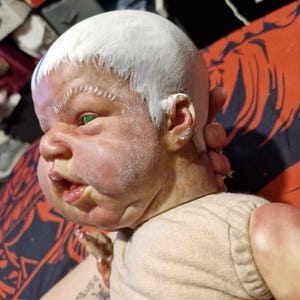 May include: A close-up of a doll's face with white paint on its head and green eyes. The doll is wearing a light beige shirt. The doll's face is painted with a realistic expression. The doll's ears have small, colorful earrings.