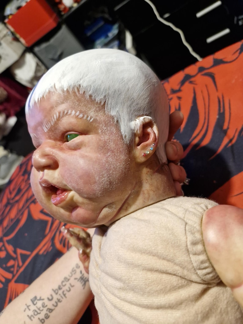 May include: A close-up of a doll's face with white paint on its head and green eyes. The doll is wearing a light beige shirt. The doll's face is painted with a realistic expression. The doll's ears have small, colorful earrings.