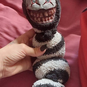 May include: A creepy sock puppet with a black and white striped body and a white face with red eyes and a wide, toothy grin.