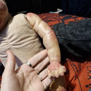 May include: Close-up of a realistic baby doll's arm and hand. The doll's skin is painted with a realistic flesh tone and has visible veins. The doll's hand is posed in a relaxed position with slightly curled fingers.