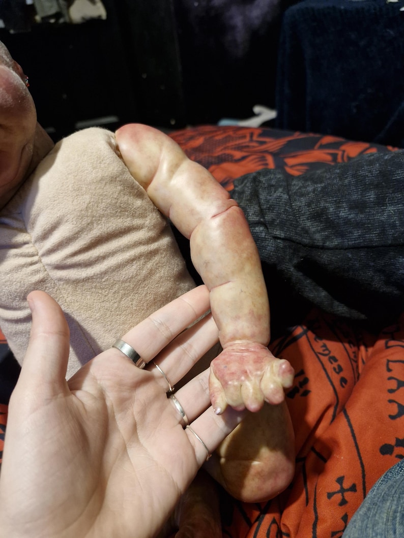 May include: Close-up of a realistic baby doll's arm and hand. The doll's skin is painted with a realistic flesh tone and has visible veins. The doll's hand is posed in a relaxed position with slightly curled fingers.