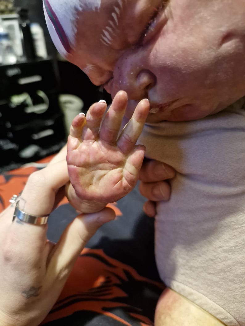 May include: A close-up of a baby doll's hand, with a person's hand holding the doll's hand. The doll's hand is painted with a realistic skin tone and has small, detailed fingernails. The doll's hand is slightly curled, as if the doll is reaching out to touch something.