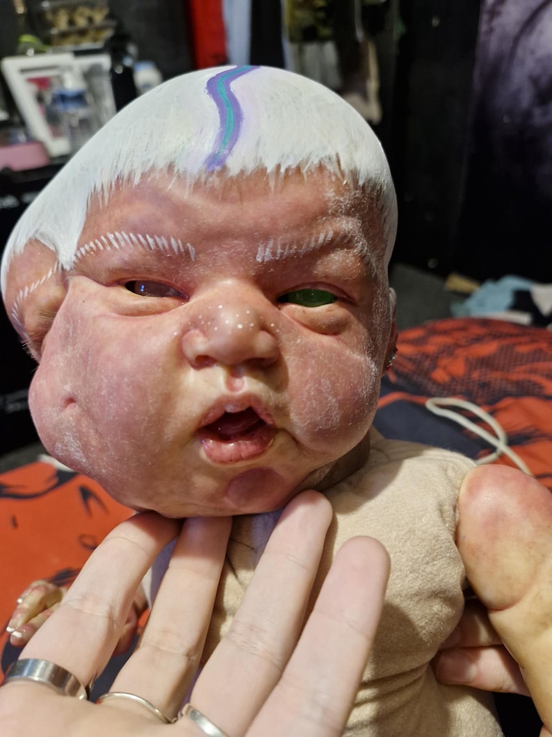May include: A realistic baby doll with a white painted head and a green eye. The doll is wearing a beige onesie and has a painted blue and purple stripe on its forehead.