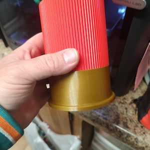 Puede incluir: Un vaso de chupito impreso en 3D de color rojo y dorado con un diseño acanalado. El vaso está siendo sostenido por una mano.