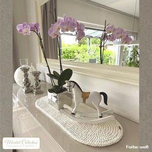 Könnte beinhalten: Ein weißes Schaukelpferd mit einem beigen Sattel steht auf einer weißen, ovalen, gehäkelten Matte. Eine Topf-Orchidee mit hellvioletten Blüten und silbernen Kerzenhaltern befinden sich ebenfalls auf der weißen Oberfläche.