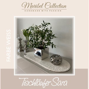 Könnte beinhalten: Ein weißer gehäkelter Tischläufer mit einem silbernen Topf und einer Pflanze auf einer hellbraunen Oberfläche. Der Text "Tischläufer Sara" ist unter dem Läufer geschrieben. Der Text "FARBE WEISS" ist auf der linken Seite des Bildes geschrieben. Der Text "Maribel Collection HANDMADE WITH PASSION" ist oben im Bild geschrieben.