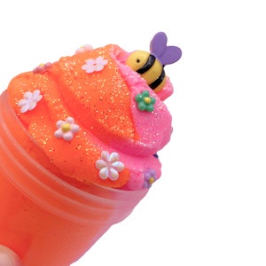 K&ouml;nnte beinhalten: Orangefarbener und rosafarbener Schleim in einem durchsichtigen Plastikbecher mit Bienen- und Blumenverzierungen.
